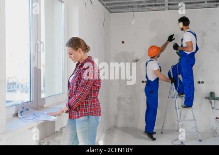 Ein Kunde steht am Fenster und untersucht das Diagramm des Hauses. Zwei Bauherren bohren eine Wand, während sie auf einer Trittleiter stehen Stockfoto