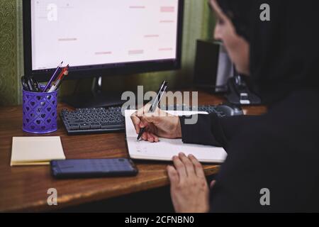 Unternehmerin aus dem Nahen Osten. Beschäftigte arabische Geschäftsfrau. Eine Frau mittleren Alters in traditioneller arabischer Kleidung, Hijab oder Abaya, die beim Schreiben arbeitet Stockfoto