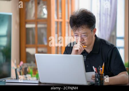 Asiatischer Mann mittleren Alters, der zu Hause arbeitet. Telearbeitkonzept. Stockfoto