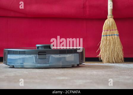 Roboter-Staubsauger und Besen auf Teppichboden gegen Rot Sofa Stockfoto