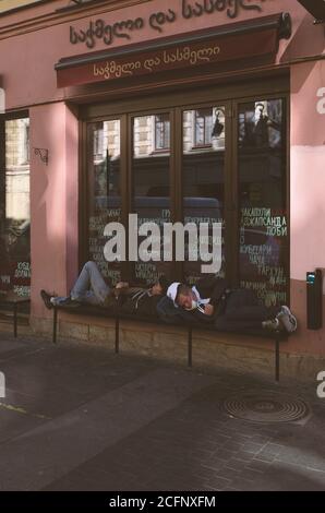Sankt Petersburg, Russland - 21. August 2020: Obdachlose schlafen auf einer Bank in der Nähe eines georgianischen Café. Das Fenster hat Inschriften in Russisch und Geor Stockfoto
