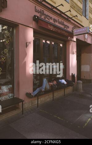 Sankt Petersburg, Russland - 21. August 2020: Obdachlose schlafen auf einer Bank in der Nähe eines georgianischen Café. Das Fenster hat Inschriften in Russisch und Geor Stockfoto