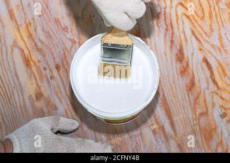 Ein Mann in Arbeitshandschuhe malt die hölzerne Oberfläche mit der weißen Farbe, legt den Pinsel in den Krug der Farbe Stockfoto