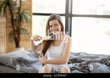 Junge Frau putzt ihre langen Haare auf dem Bett, nachdem sie morgens aufgewacht ist, drinnen Stockfoto
