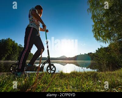 Sport Körper Junge bleiben auf Push Roller am Rand am Teich Wehr. Der Sonnenuntergang über dem gegenüberliegenden Ufer spiegelt sich im Wasserspiegel des Teichs. Stockfoto