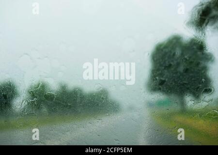 Regen auf einer Windschutzscheibe eines Autos mit verschwommener Straße Und bewölktem Himmel im Hintergrund Stockfoto