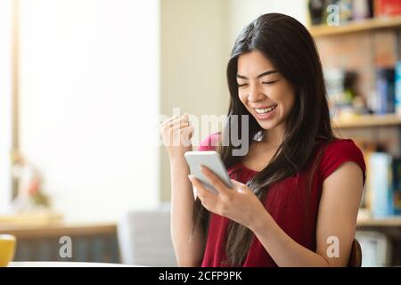 Überglücklich asiatische Frau feiert Erfolg mit Smartphone im Café, erhielt gute Nachrichten Stockfoto