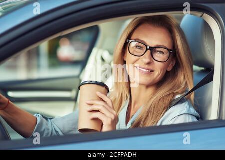 Porträt einer lächelnden reifen Frau mit Brillen, die hinter dem Lenkrad ihres modernen Autos sitzt, eine Tasse Kaffee zum Mitnehmen hält und anschaut Stockfoto
