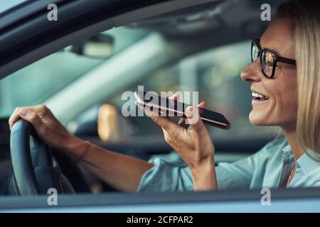 Seitenansicht einer lächelnden Geschäftsfrau mit Smartphone, die während der Fahrt Sprachnachrichten aufzeichnet Stockfoto