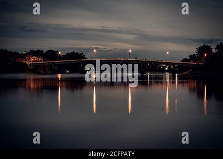 Langzeitaufnahme der alten Eisenbahnbrücke über den Fluss bei Nacht mit Besinnung in Ratchaburi Thailand Stockfoto