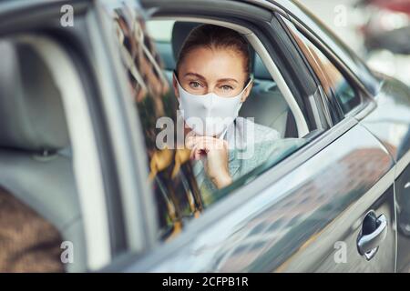 Kaukasische Geschäftsfrau trägt medizinische Schutzmaske auf dem Rücksitz sitzen Im Auto und aus dem Fenster schauen Stockfoto