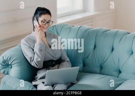 Zufrieden Geschäftsfrau in eleganter Kleidung diskutiert finanzielle Fragen mit Partner über Handy, Posen auf bequemen Sofa, sitzt auf modernen Laptop Co Stockfoto