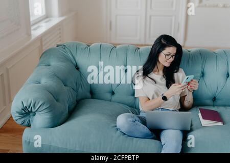 Blick von oben auf beschäftigt Freiberuflerin sitzt in gemütlichen Raum auf bequemen Sofa, arbeitet an neuen Startup-Projekt, nutzt Laptop-Computer, hält mobile phon Stockfoto