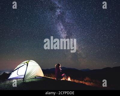 Weibliche Camper mit einer Pause in der Sommernacht Camping in den Bergen neben Lagerfeuer, glühenden touristischen Zelt. Junge Frau genießen Blick auf den Nachthimmel voller Sterne und Milchstraße. Tourismus Abenteuer Konzept Stockfoto