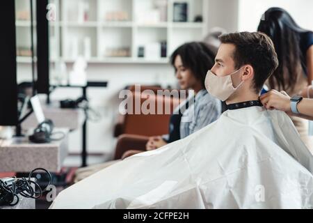 Junger Mann in Schutzmaske sitzt in Client Stuhl im Friseurladen, warten auf Haarschnitt Stockfoto