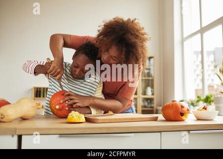 Stilvolle afroamerikanische Frau verbringt Zeit mit ihrem Sohn stehen Am Tisch Kürbis für Halloween mit Küchenmesser zusammen schnitzen Stockfoto