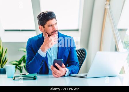 Ein denkender Geschäftsmann, der am Büroschreibtisch sitzt und sein Mobiltelefon benutzt, während er SMS-Nachrichten abgibt. Stockfoto