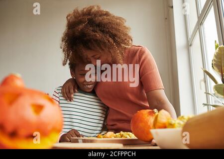 Liebevolle Mutter umarmt ihren fröhlichen Sohn, während sie reife Kürbisse schnitzt Für Halloween Party zusammen Stockfoto