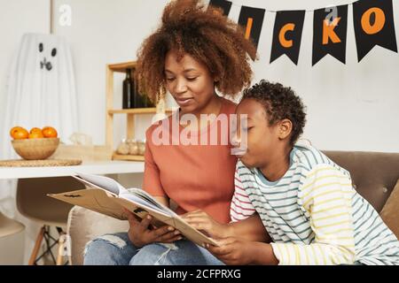 Schöne afroamerikanische Frau sitzt auf dem Sofa im Wohnzimmer Mit ihrem präteen Sohn Lesung interessante Halloween Geschichten Buch Stockfoto