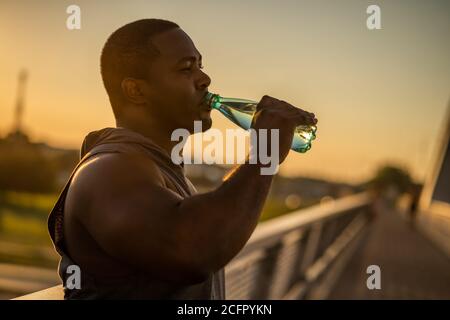 Der junge afroamerikanische Mann trinkt nach dem Training auf der Brücke in der Stadt Wasser. Stockfoto
