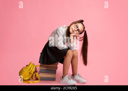 Schulmädchen in Brille mit Pferdeschwänzen und Rucksack sitzt auf Stapel Von Büchern und schaut auf leeren Raum Stockfoto