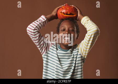 Fröhlicher afroamerikanischer Junge in gestreiftem Langarm-Hemd mit Jack O' Lantern auf dem Kopf lächelnd auf Kamera, Studioportrait Stockfoto