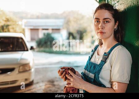 Gleichstellung der Geschlechter. Ein Porträt einer jungen Brünette in Uniform, die sich gegen das Tor lehnt und ihre Hände mit einem Lappen abwischt. Im Hintergrund eine Straße und ein Stockfoto