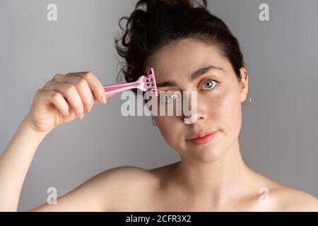 Eine schöne junge Frau hält einen Rasierer an ihre Augenbraue. Das Konzept der loszuwerden unerwünschte Gesichtshaar. Stockfoto