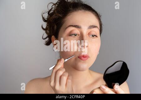 Eine junge hübsche Frau schaut in einen kompakten Spiegel und zieht ihre Antennen mit einer Pinzette heraus. Das Konzept der loszuwerden unerwünschte Haare. Stockfoto