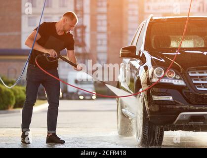 Der junge Trendmann wäscht die Räder seines Autos draußen und hält einen Schlauch mit einem Wasserstrahl unter hohem Druck, voller Länge Stockfoto