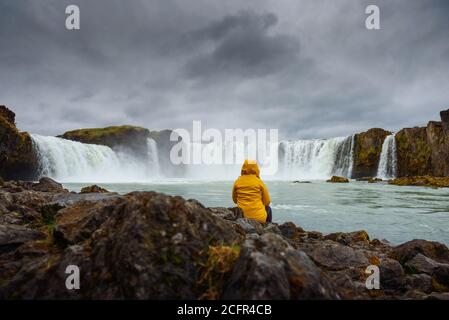 Tourist in einer gelben Jacke, entspannend am Wasserfall Godafoss in Island Stockfoto