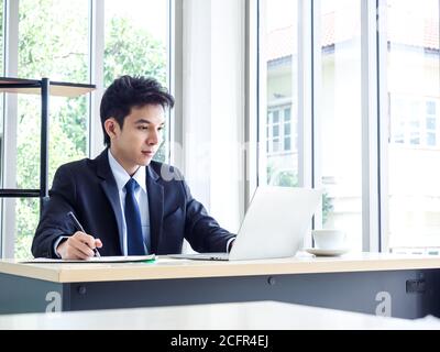 Junge asiatische Geschäftsmann im Anzug mit Laptop-Computer im Büro arbeiten. Mann in Anzug mit Laptop an gut beleuchteten Arbeitsplatz. Stockfoto