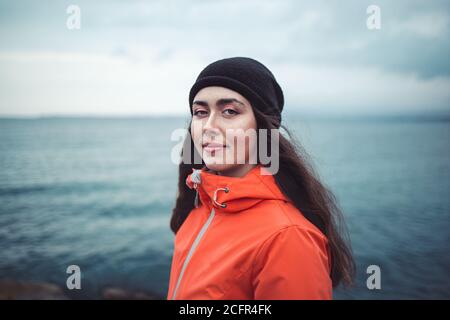 Porträt einer lächelnden Brünette Frau mit langen Haaren, trägt einen Hut und eine orangefarbene Jacke. Im Hintergrund das Meer und die Horizontlinie. Speicherplatz kopieren. Stockfoto