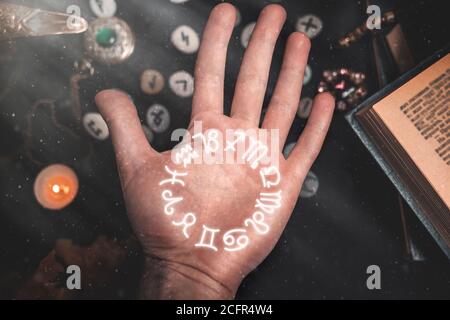 Astrologie und Esoterik. Nahaufnahme der männlichen Handfläche. Auf schwarzem Hintergrund liegen Runen, eine Kerze, Amulette und ein Buch. Der Blick von oben. Sternzeichen Horoskop Stockfoto