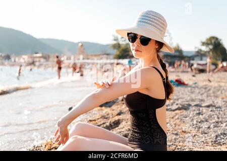 Eine Frau in einem Strohhut, Badeanzug und Brille schmiert Sonnencreme am Strand sitzen. Das Konzept der richtigen Bräunung. Stockfoto