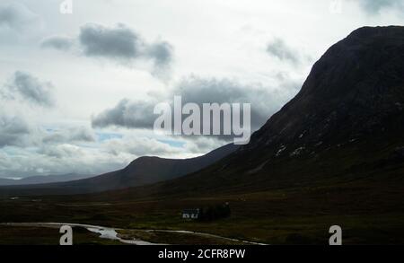 Schottische Highlands, Nationalparks - EINE einsame kleine weiße Hütte im Tal neben Bergen und einem Fluss an einem grauen stürmischen Tag in der Nähe von Glen Coe. Stockfoto