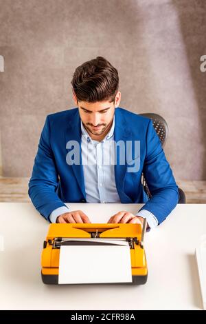 Aufnahme eines hübschen Mannes, der am Schreibtisch sitzt und eine Retro-Schreibmaschine benutzt. Stockfoto