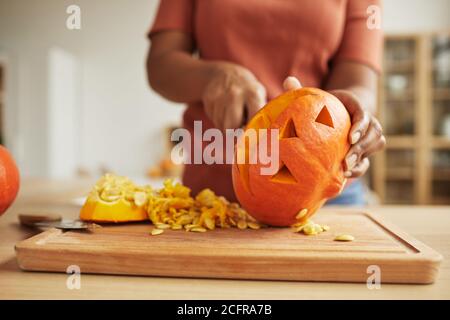 Nicht erkennbare afroamerikanische Frau, die am Tisch steht und Jack-O'-Laterne herausschnitzt Von reifen orangefarbenen Kürbis Stockfoto