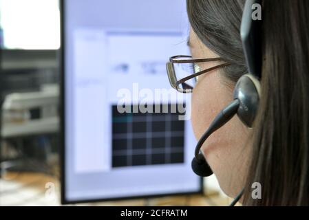 Straffes Porträt einer jungen Frau mit Brille, von hinten betrachtet, mit einem Headset und Mikrofon vor ihrem Computerbildschirm Stockfoto