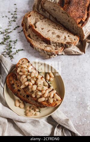 Gekochte Cannellini-Bohnen auf geröstetem Sauerteigbrot mit frischem Thymian und einem in Scheiben geschnittenen Laib Brot. Stockfoto