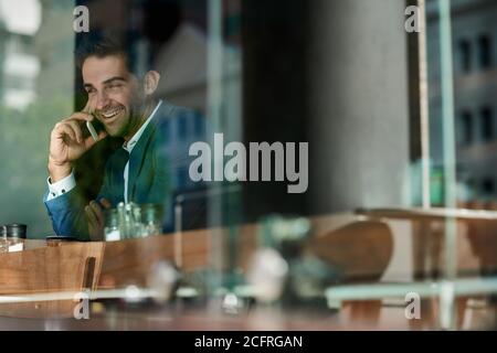 Lächelnder Geschäftsmann, der sich an einem Cafe-Fenster auf seinem Handy unterhielt Stockfoto