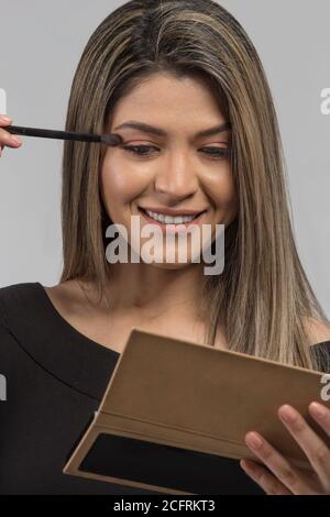 Gesicht der lateinischen Frau mit blonden und langen Haaren auf Make-up setzen mit der anderen Hand hält einen Spiegel, Studio Stockfoto