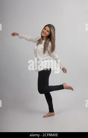 Junge schlanke Frau springt mit einem Fuß in die Luft, trägt weiße Bluse und schwarze Hose, Studio mit grauem Hintergrund Stockfoto