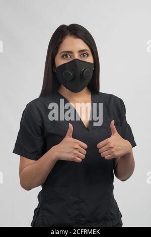 Frau in schwarzer Uniform mit maskhis Hände vorne und Daumen nach oben, Studio gekleidet Stockfoto