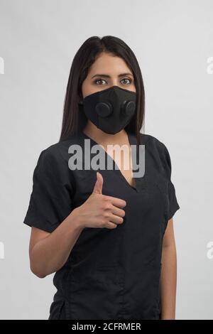 Frau in schwarzer Uniform mit einer Maske über ihrer Mundhand mit Daumen nach oben, studiowith grauen Hintergrund gekleidet Stockfoto