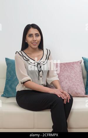 Junge lateinerin mit schwarzen Haaren sitzt auf einem weißen Sofa mit bunten Kissen, trägt eine weiße Bluse und schwarze Hose Stockfoto