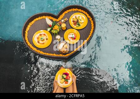 Schwimmende Frühstück im Pool. Frau Hände halten Teller mit frischen Früchten. Sommer gesunde Ernährung. Tropischer Lebensstil. Rattan Tablett, orange Blumen, Bali Stil und Service. Stockfoto