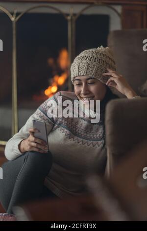 Die junge Lateinerin sitzt mit einem Handy, das ihre Hand hält und ihren Kopf berührt, trägt einen Wollhut, eine farbige Jacke und eine schwarze Hose Stockfoto