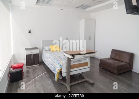 Krankenhauszimmer mit Liege, beweglichem Tisch, gelbem Kissen, weißen Wänden, Holzboden, Kaffeesofa und Regalen Stockfoto