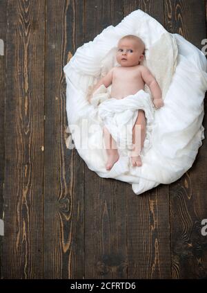 Kleinkind mit weißer Decke bedeckt. Baby liegt auf weißen weichen Bettdecke als Wolke oder Nest entworfen. Kindheit und Göttliche Geschenk Konzept. Neugeborenes Kleinkind mit blauen Augen und interessiertes Gesicht auf Holzhintergrund Stockfoto